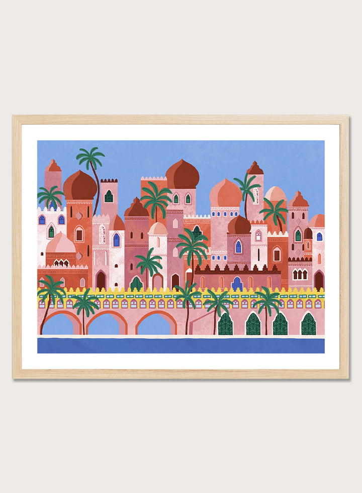 A framed art print featuring a stylized illustration of a coastal city with pink and peach buildings, green palm trees, and a blue sky. Arabian Fantasy City by Ceyda Alasar travel art print featuring ornate domes, arches, palm trees, patterned facades, and colorful desert architecture.