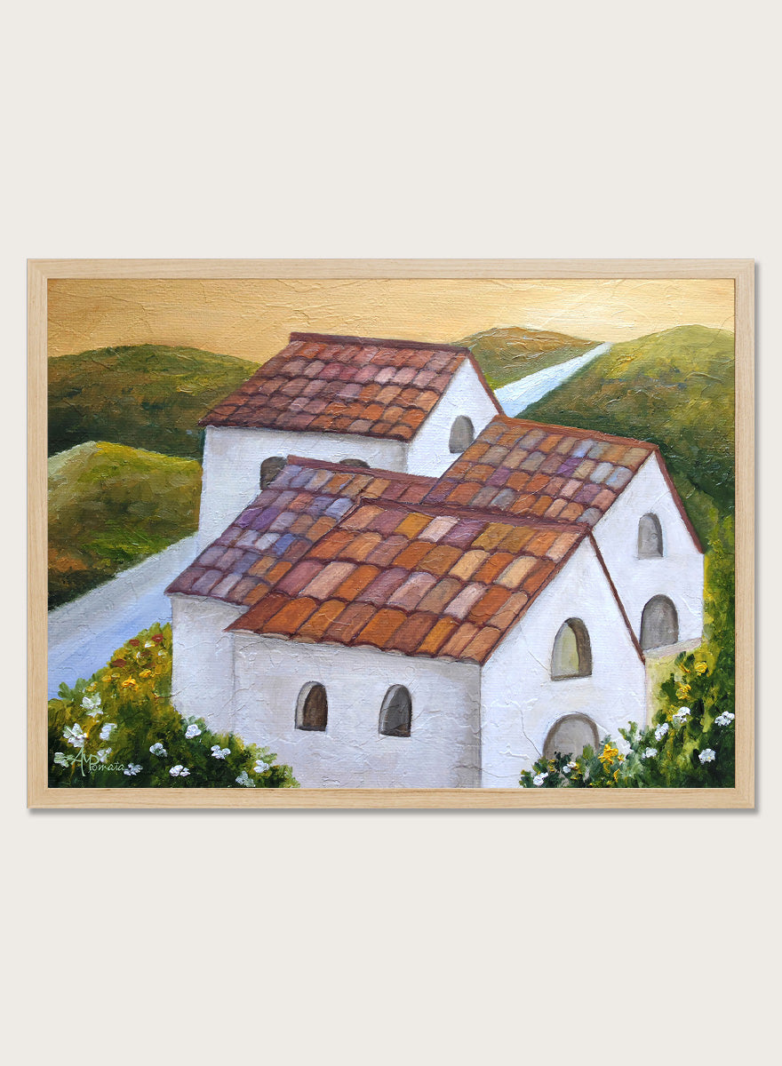 A framed landscape painting depicting two white buildings with terracotta tiled roofs nestled in green hills, with a winding light blue road and a warm yellow sky. Shop Tuscan Hillside Village by Angeles M. Pomata, a warm countryside landscape art print featuring white houses, terracotta roofs, green hills, and wildflowers.