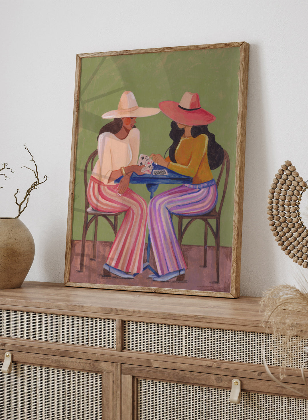 Two women playing cards at a table with colorful clothing and hats against a green background. Stylish Women Play Cards by Caroline Bonne Müller is a fine art print and wall poster featuring two fashionable women playing cards at a table.