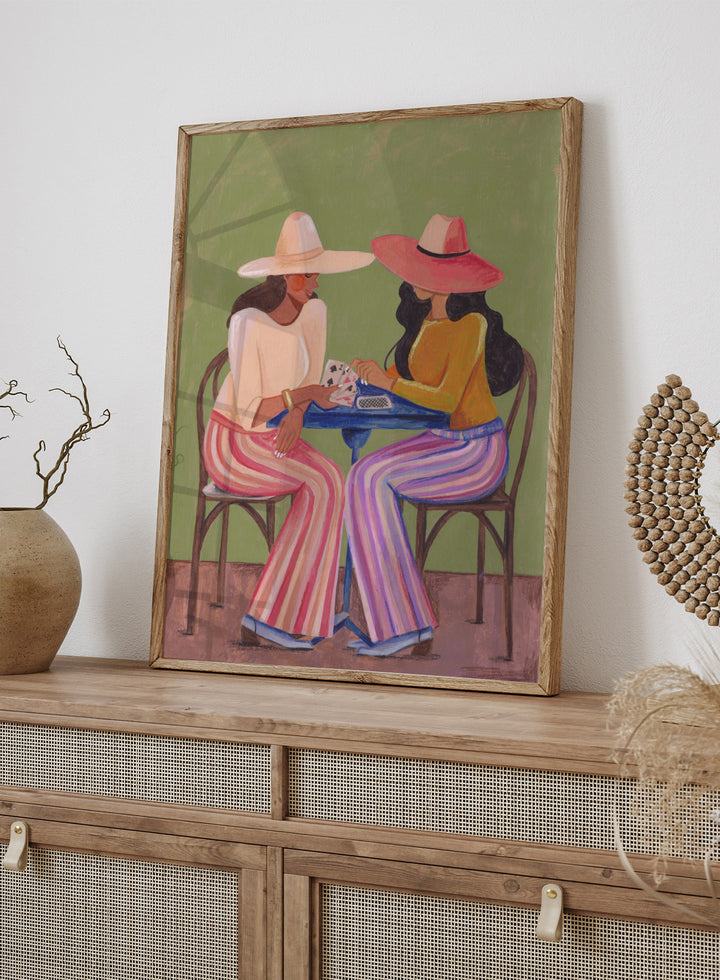 Two women playing cards at a table with colorful clothing and hats against a green background. Stylish Women Play Cards by Caroline Bonne Müller is a fine art print and wall poster featuring two fashionable women playing cards at a table.