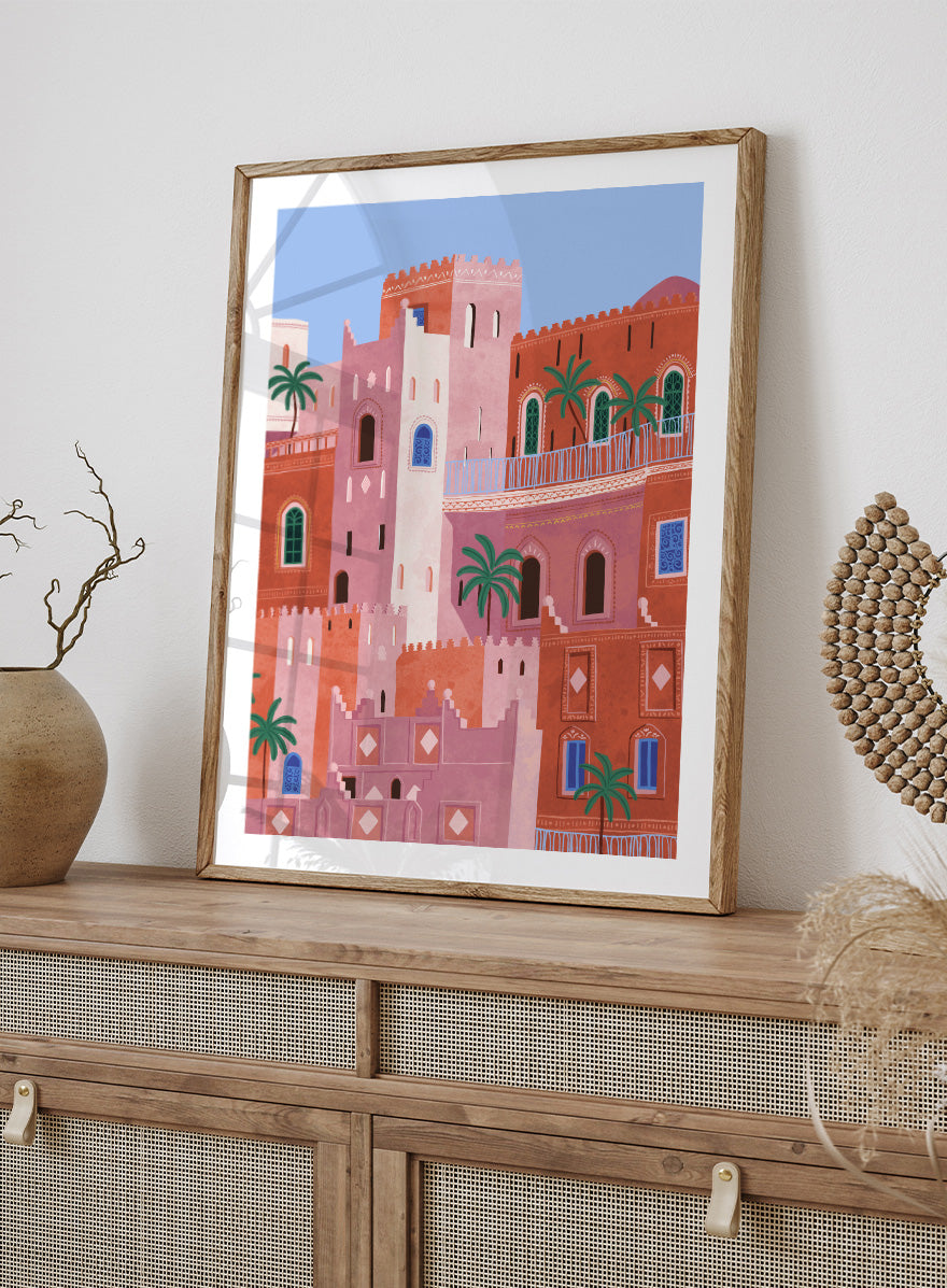 A framed art print featuring a stylized illustration of Moroccan-style buildings in shades of terracotta, pink, and cream, accented with green palm trees and blue window details, set against a light blue sky. Vibrant Moroccan City by Ceyda Alasar travel art print featuring layered terracotta architecture, arched windows, palms, and colorful Moroccan details.