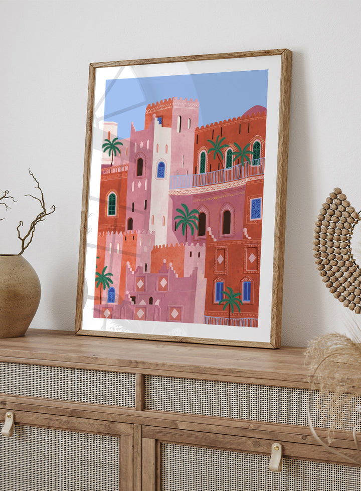 A framed art print featuring a stylized illustration of Moroccan-style buildings in shades of terracotta, pink, and cream, accented with green palm trees and blue window details, set against a light blue sky. Vibrant Moroccan City by Ceyda Alasar travel art print featuring layered terracotta architecture, arched windows, palms, and colorful Moroccan details.