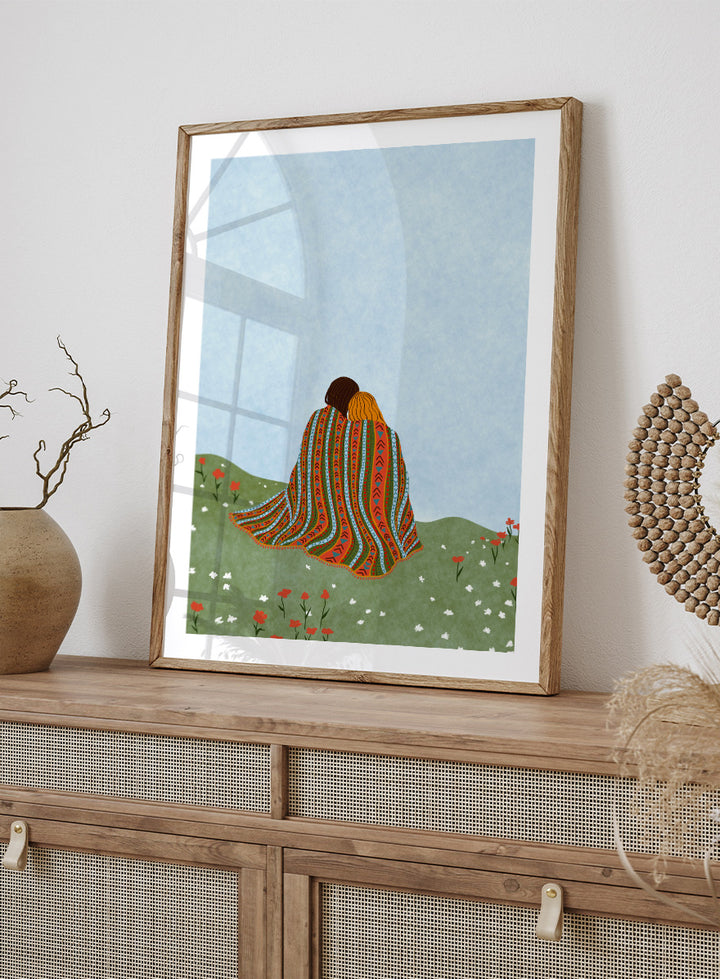 A framed art print featuring an illustration of two people sitting on a grassy hill, wrapped in a vibrant, patterned blanket under a light blue sky. Cozy Hillside Embrace by Ceyda Alasar art print featuring two people wrapped in a patterned blanket among green hills and wildflowers.