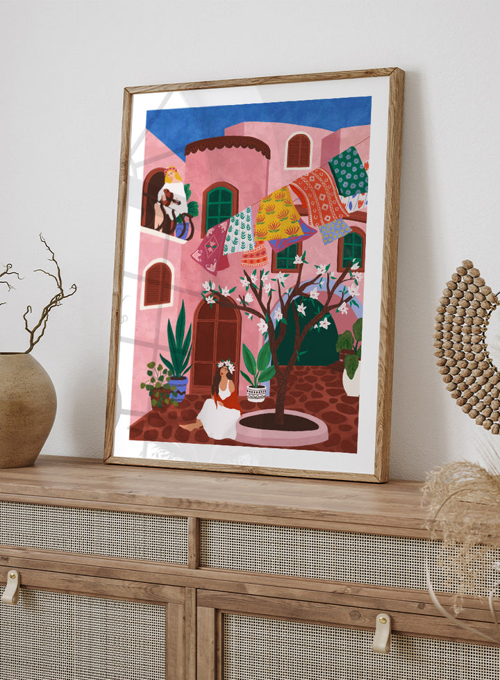 A framed art print featuring a colorful Mediterranean-style illustration of a pink courtyard with a woman sitting under a flowering tree, laundry hanging in the background, and a blue sky above. Pink Village Courtyard by Ceyda Alasar art print featuring pink architecture, patterned rugs, flowering trees, potted plants, and relaxed courtyard life.