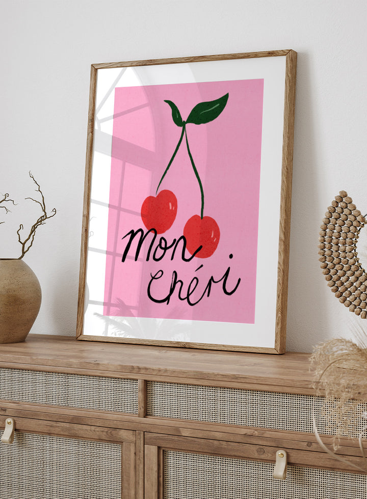 Two red cherries with green stems on a pink background and 'mon chéri' text. Mon Chéri by Dirkie Nell is a cherry typography art print featuring bold red fruit and playful lettering. A colorful love-inspired wall poster.