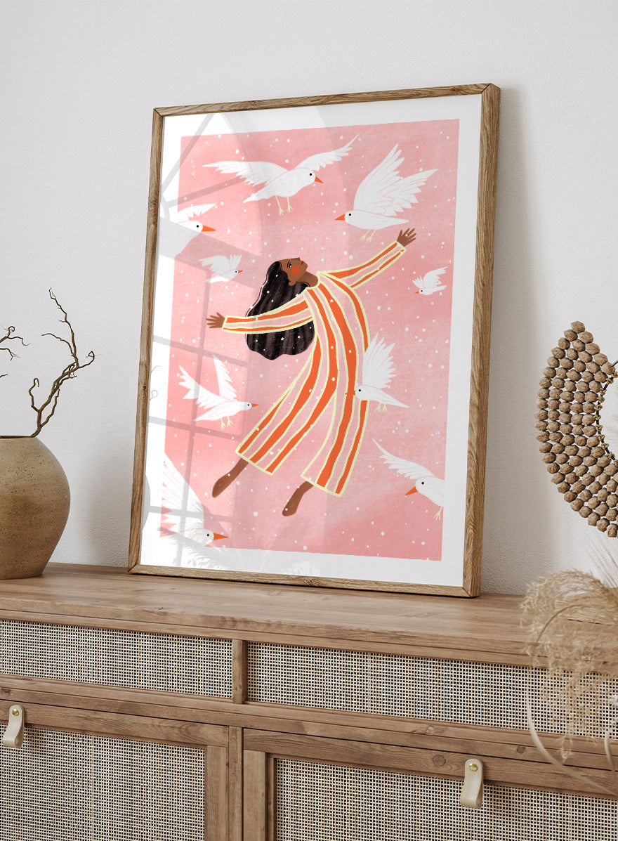 A framed art print featuring an illustration of a woman in orange and white striped pajamas floating among white doves on a soft pink background. Joyful Liberation by Bea Müller woman art print featuring a free-spirited figure surrounded by white birds in bold pink and orange tones.