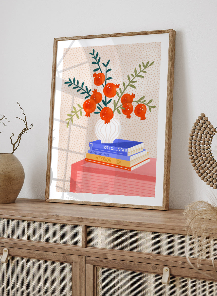 A framed art print featuring a stylized illustration of pomegranate branches in a white vase resting on a stack of books, set against a light beige dotted background. Cozy Tabletop Pomegranates by Bea Müller still life art print featuring red fruit branches, a sculptural vase, design books, and colorful interior styling.