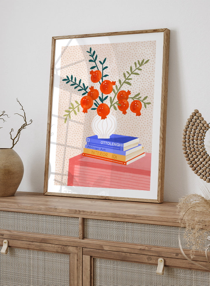 A framed art print featuring a stylized illustration of pomegranate branches in a white vase resting on a stack of books, set against a light beige dotted background. Cozy Tabletop Pomegranates by Bea Müller still life art print featuring red fruit branches, a sculptural vase, design books, and colorful interior styling.