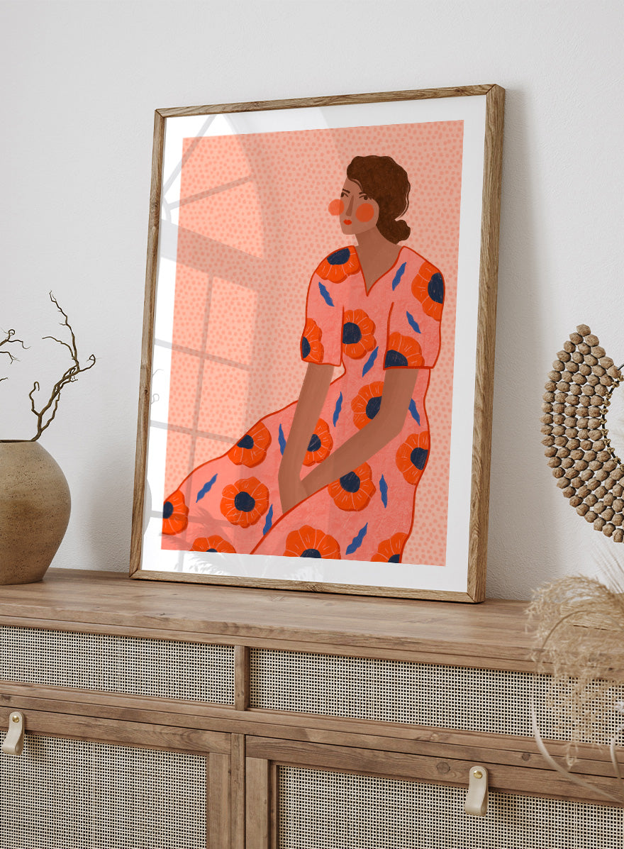 A stylized fashion illustration of a seated woman in a coral pink dress with large orange poppy motifs and blue leaf accents, shown with a white border and black frame. Stylish Poppy Portrait by Bea Müller floral woman art print featuring a pink dress covered in orange poppies and blue leaves for colorful modern wall decor.