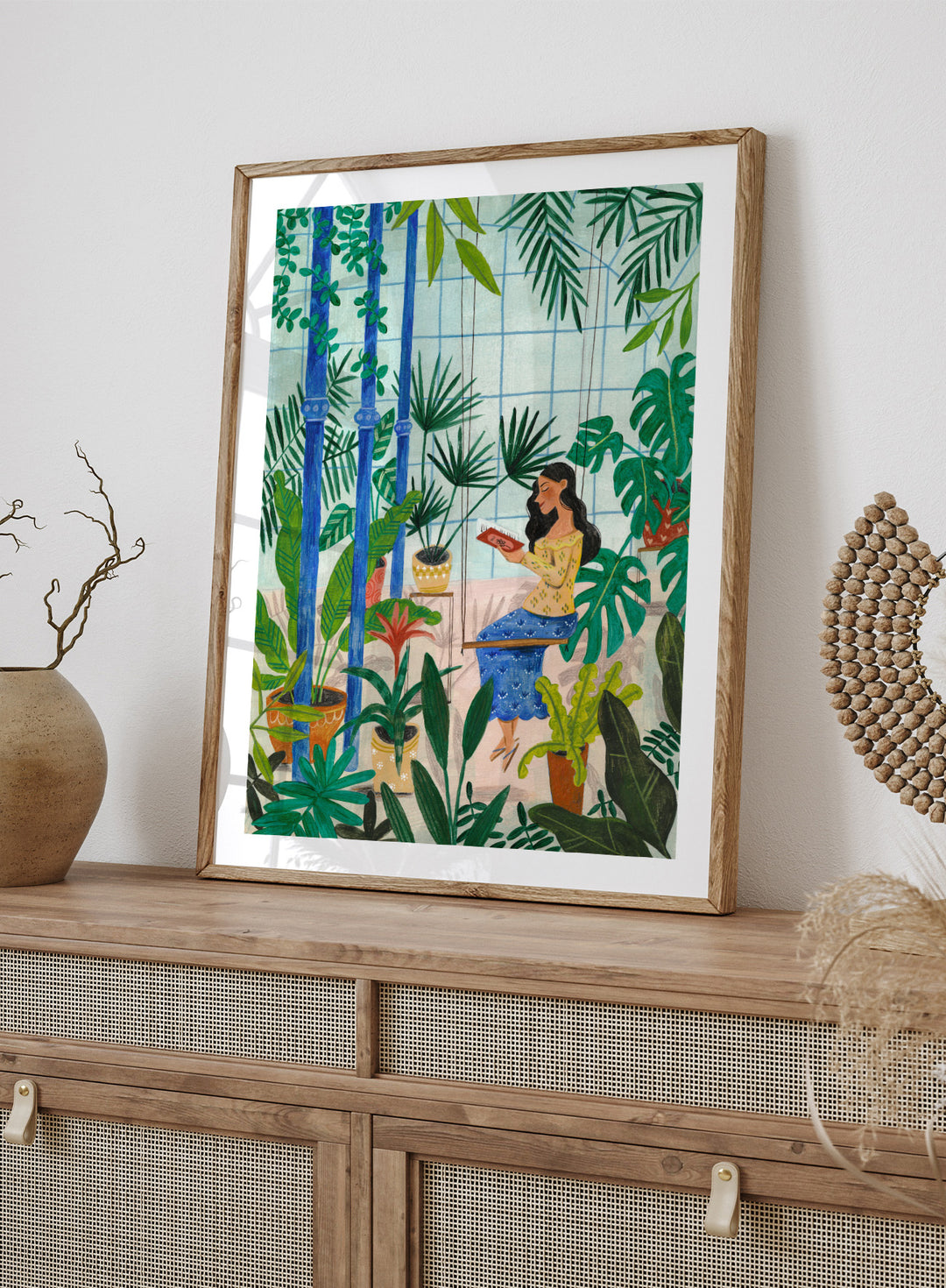 Woman sitting among plants in a room with bamboo and various greenery. Botanical Greenhouse Reader by Caroline Bonne Müller is a fine art print and wall poster featuring a woman reading in a lush indoor garden filled with tropical plants.