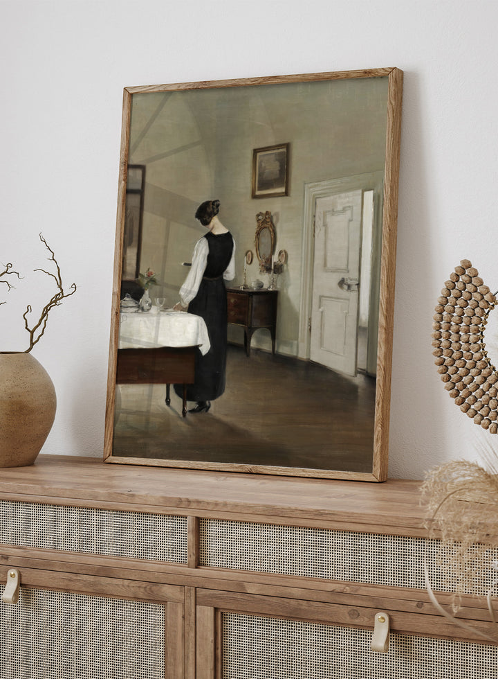 Woman in a black dress standing in a room with a table and mirror. In the Dining Room by Carl Holsøe is a vintage Danish interior art print featuring a woman in a softly lit dining room with muted tones and classic Scandinavian style.