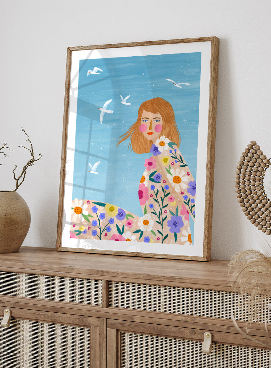 A framed art print featuring a stylized illustration of a young woman with long light brown hair, surrounded by colorful flowers and white birds against a light blue sky. ree Spirit Floral Dreams by Bea Müller floral woman art print featuring a flowing flower-covered summer dress, white birds, and a bright blue sky.
