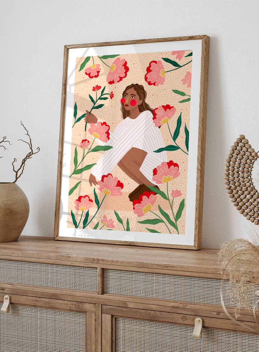 A framed art print featuring a stylized illustration of a woman in a white striped sweater surrounded by pink and red flowers on a peach background. Woman in Floral Garden by Bea Müller floral art print featuring a woman surrounded by oversized pink flowers, green leaves, and playful botanical details.
