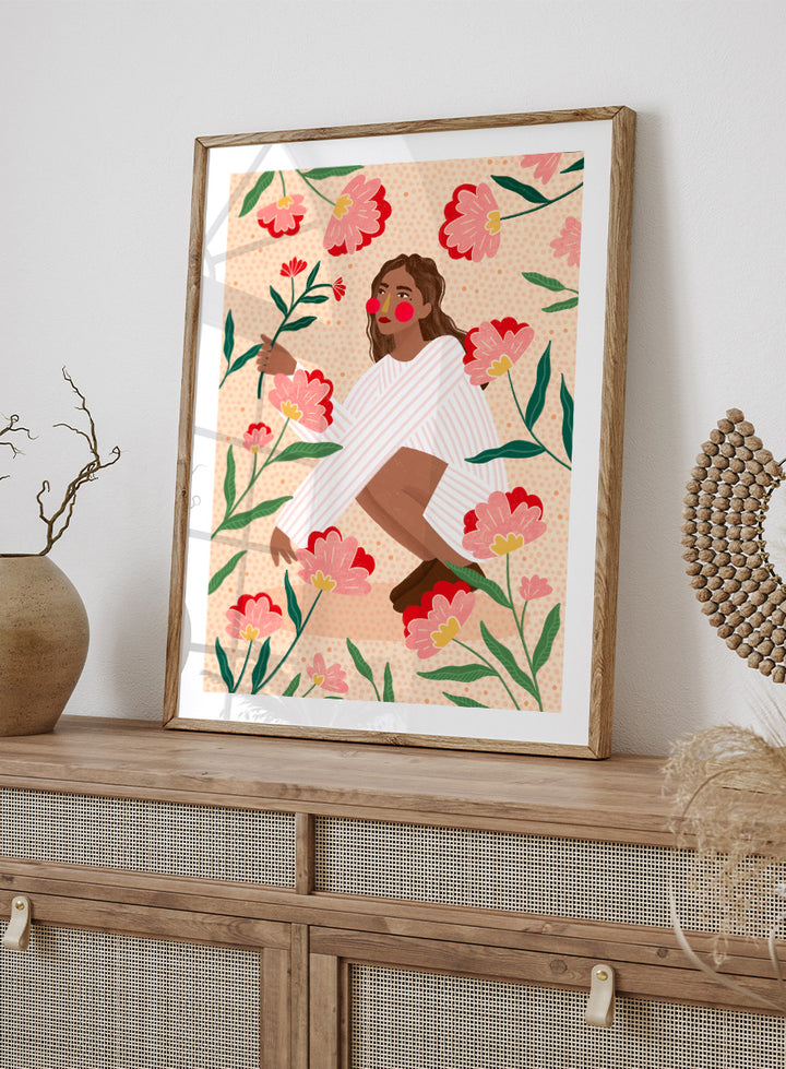 A framed art print featuring a stylized illustration of a woman in a white striped sweater surrounded by pink and red flowers on a peach background. Woman in Floral Garden by Bea Müller floral art print featuring a woman surrounded by oversized pink flowers, green leaves, and playful botanical details.