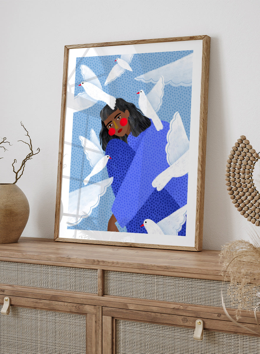 A framed art print featuring a stylized illustration of a woman with dark hair and red cheeks, wrapped in a blue patterned blanket, surrounded by white doves against a light blue dotted background. Peaceful Woman and Doves by Bea Müller art print featuring a woman in blue surrounded by white birds, layered textures, and soft modern color.