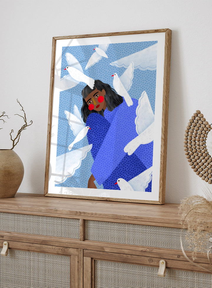 A framed art print featuring a stylized illustration of a woman with dark hair and red cheeks, wrapped in a blue patterned blanket, surrounded by white doves against a light blue dotted background. Peaceful Woman and Doves by Bea Müller art print featuring a woman in blue surrounded by white birds, layered textures, and soft modern color.