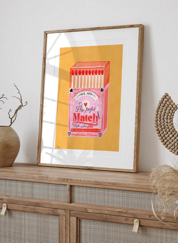 Illustration of a matchbox with 'We are the perfect match' text on a yellow background. Match Box, The Perfect Match by Baroo Bloom is a playful love-themed art print featuring a pink illustrated matchbox. Officially licensed wall poster with optional framing.