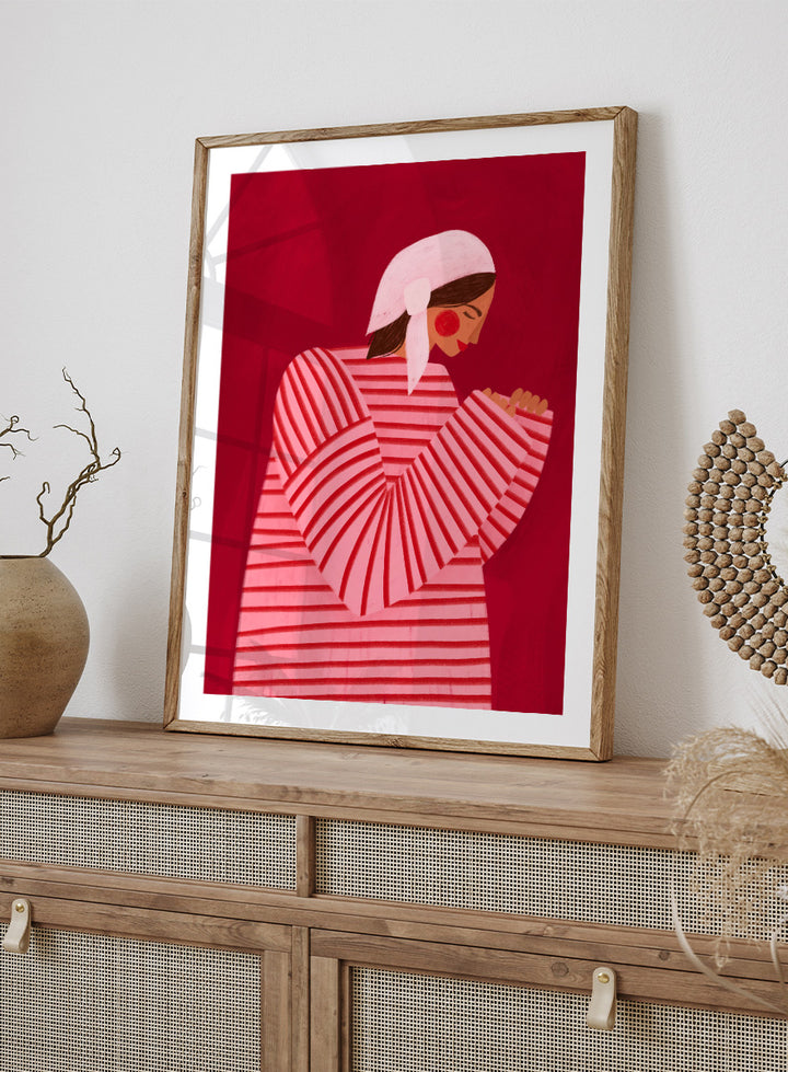 A framed art print featuring a stylized illustration of a woman in a pink and red striped top and white headscarf against a deep red background. Serene Praying Woman by Bea Müller portrait art print featuring a reflective woman in a pink striped dress against a rich red background.