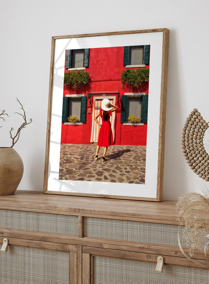 Woman in a red dress and white hat standing in front of a red wall with green shutters. Red Art Print by Anette Ohlendorf featuring a woman in a red dress standing against a red building. Officially licensed travel photography with strong color and symmetry.