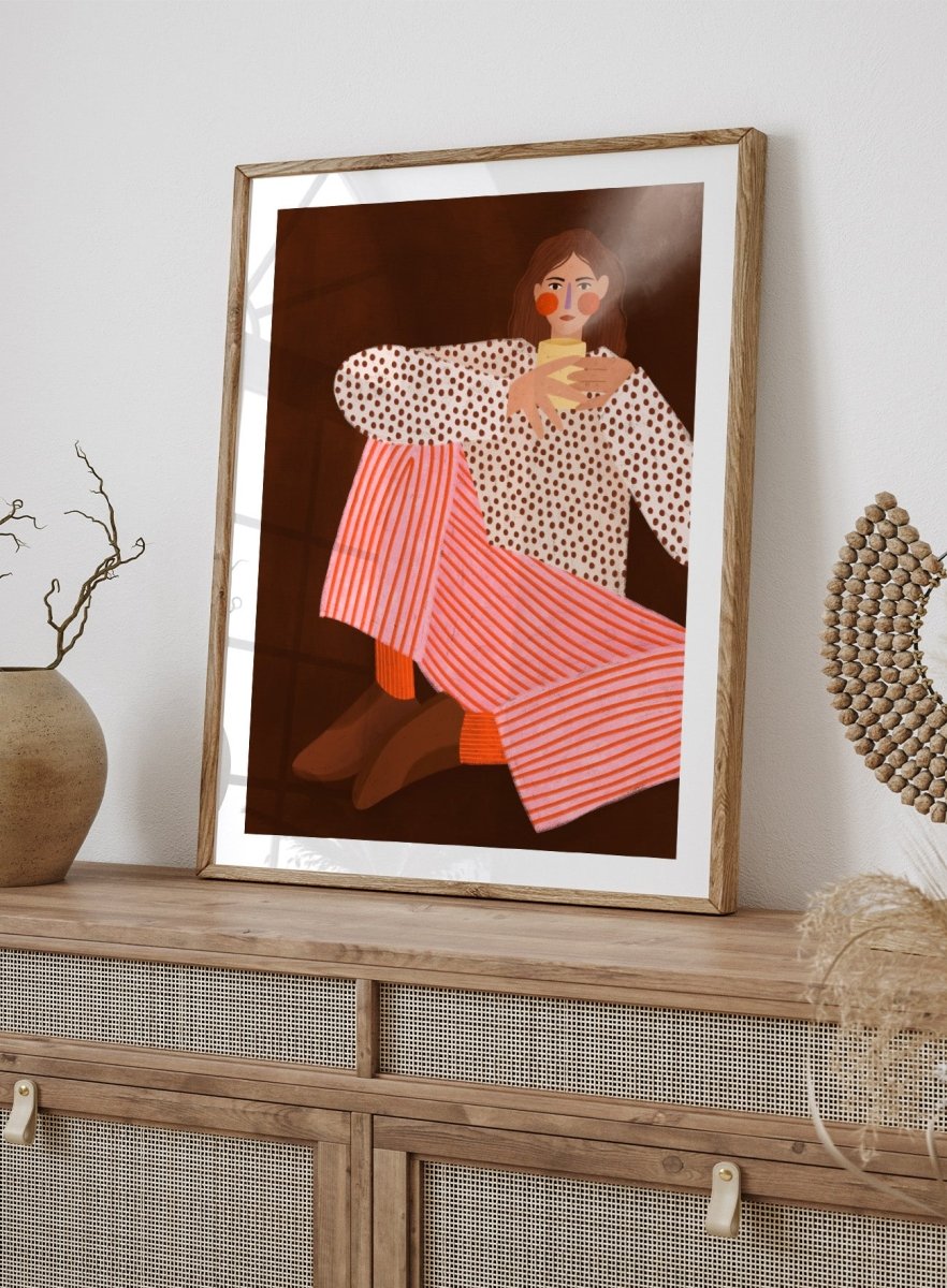 A framed art print featuring a stylized illustration of a woman in a white blouse with brown polka dots and pink-and-white striped trousers, holding a yellow cup against a dark brown background. Cozy Coffee Moment by Bea Müller premium framed fine art print featuring a woman curled up with a warm mug in bold pink, red, and brown tones.