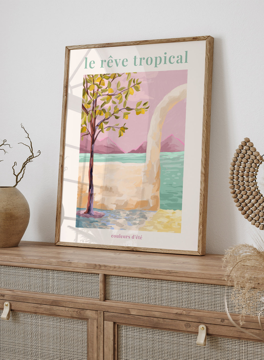 Artistic depiction of a tropical scene with a tree, beach, and mountains, featuring the text 'le rève tropical' and 'couleurs d'été'. Le Rêve Tropical Lemon Tree by Goed Blauw is a coastal art print and wall poster featuring a lemon tree, archway, and ocean view in warm pastel tones.