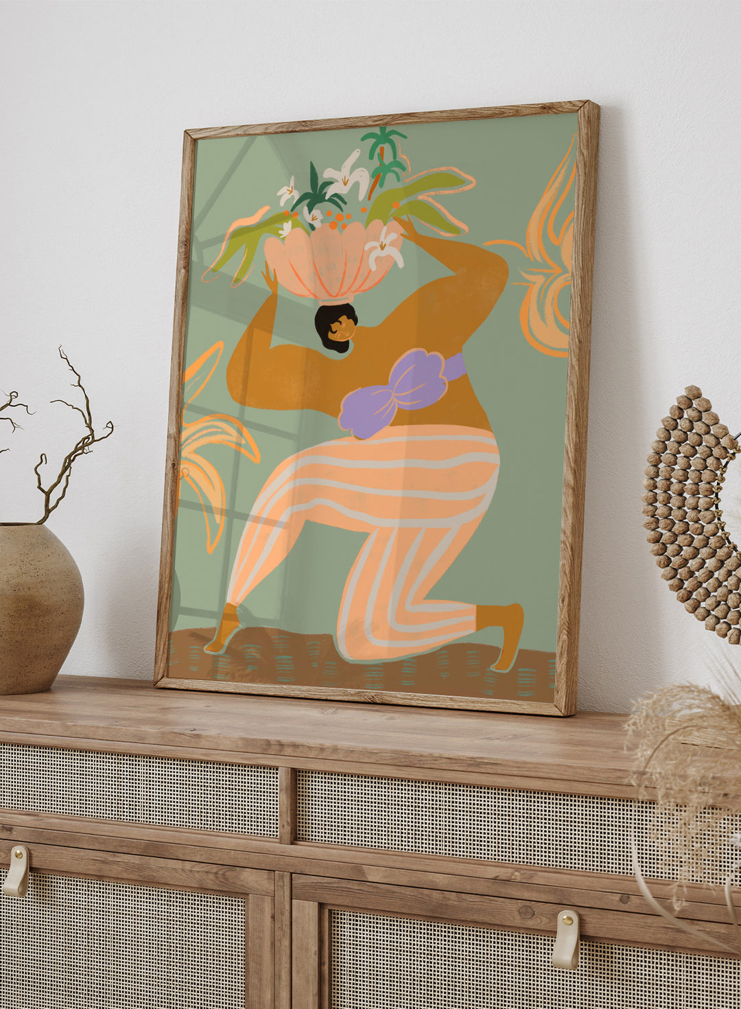 A stylized art print featuring a woman in flowing peach and white striped pants with a purple sash, carrying a large flower basket on her head against a muted sage green background. The Weight of the World by Arty Guava premium framed fine art print featuring a woman carrying tropical flowers in soft green, peach, and lilac tones.