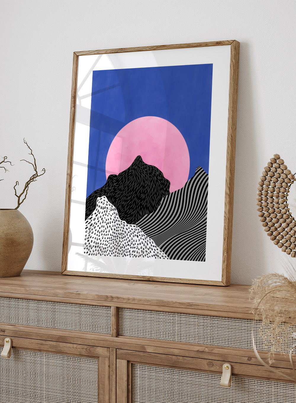Artistic depiction of mountains with a pink moon in a blue sky. Surreal Peaks View by Elena Ristova is a minimalist abstract landscape print with bold mountains and a pink sun. Officially licensed wall poster with framing.