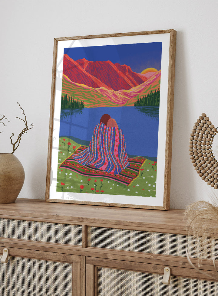 A framed art print featuring a stylized landscape illustration of two people sitting together on a patterned blanket by a blue lake, with red mountains and a yellow sun in the background. Cozy Lake Sunset art print by Ceyda Alasar features a lakeside embrace, vivid mountains, and a glowing sunset in rich, story-filled color.