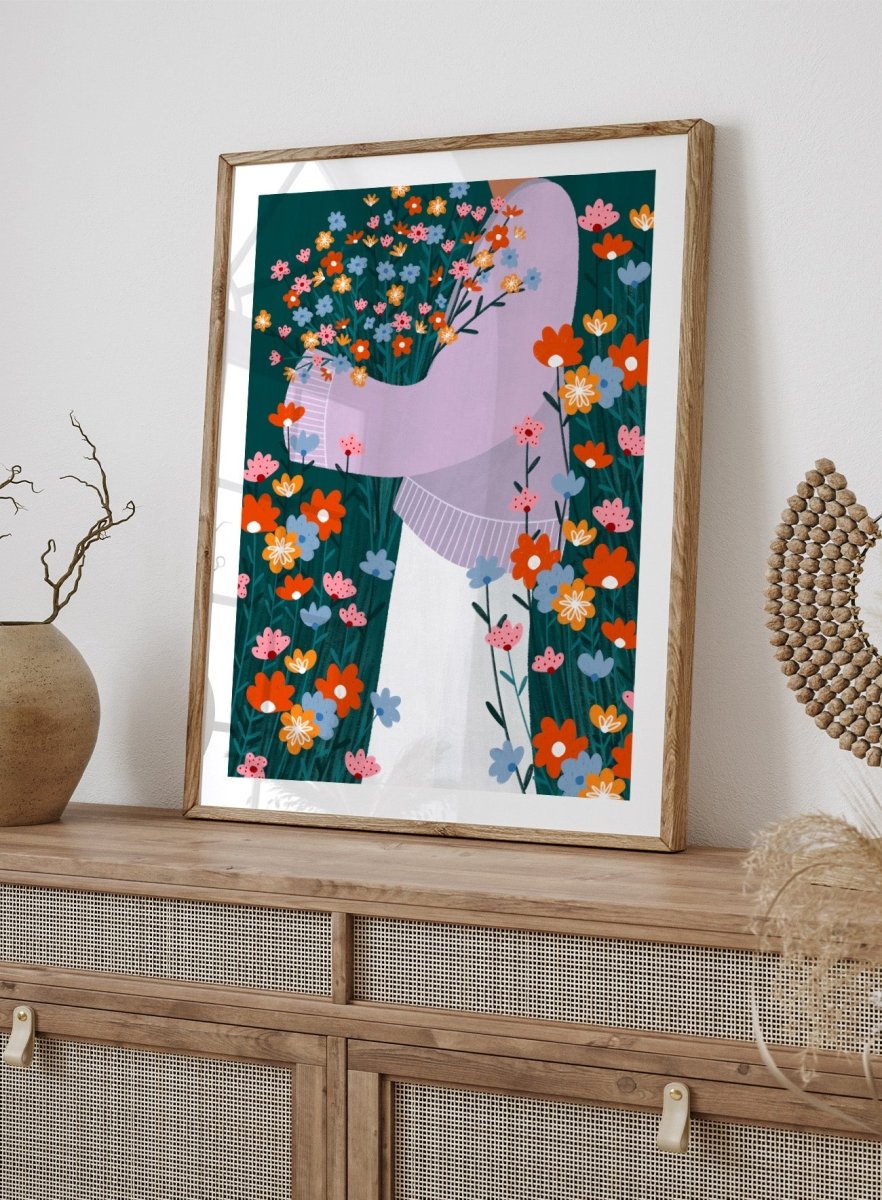 A stylized art print featuring a woman in a lavender sweater and white skirt holding a large bouquet of colorful wildflowers, framed by dense flowers on a dark teal background. Happy Woman Holding Flowers by Bea Müller premium framed fine art print featuring a woman carrying wildflowers in bright orange, pink, blue, and teal.