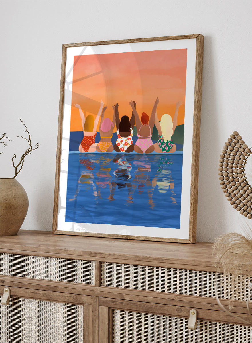 A framed art print featuring a stylized illustration of five women sitting in a pool at sunset, seen from behind as they raise their arms. The background is a warm orange sky, and the water below reflects their silhouettes. Summer Sunset Friends by Ceyda Alasar friendship art print featuring five women by the water in patterned swimsuits beneath a glowing orange sky.