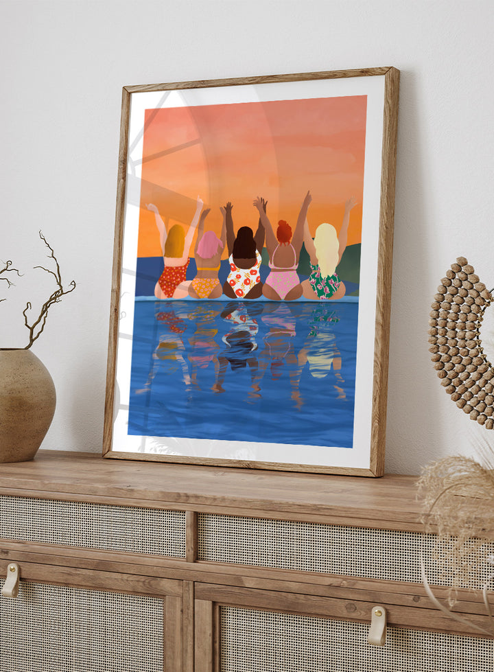 A framed art print featuring a stylized illustration of five women sitting in a pool at sunset, seen from behind as they raise their arms. The background is a warm orange sky, and the water below reflects their silhouettes. Summer Sunset Friends by Ceyda Alasar friendship art print featuring five women by the water in patterned swimsuits beneath a glowing orange sky.