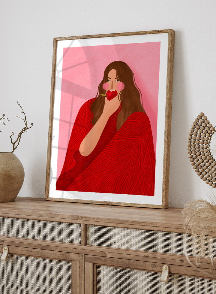 A framed art print featuring a stylized illustration of a woman with long brown hair, wearing a red patterned wrap, holding a red apple to her lips, set against a pink background. Cozy Woman Apple by Bea Müller portrait art print featuring a woman eating a red apple in bold red and pink tones for colorful modern wall decor.