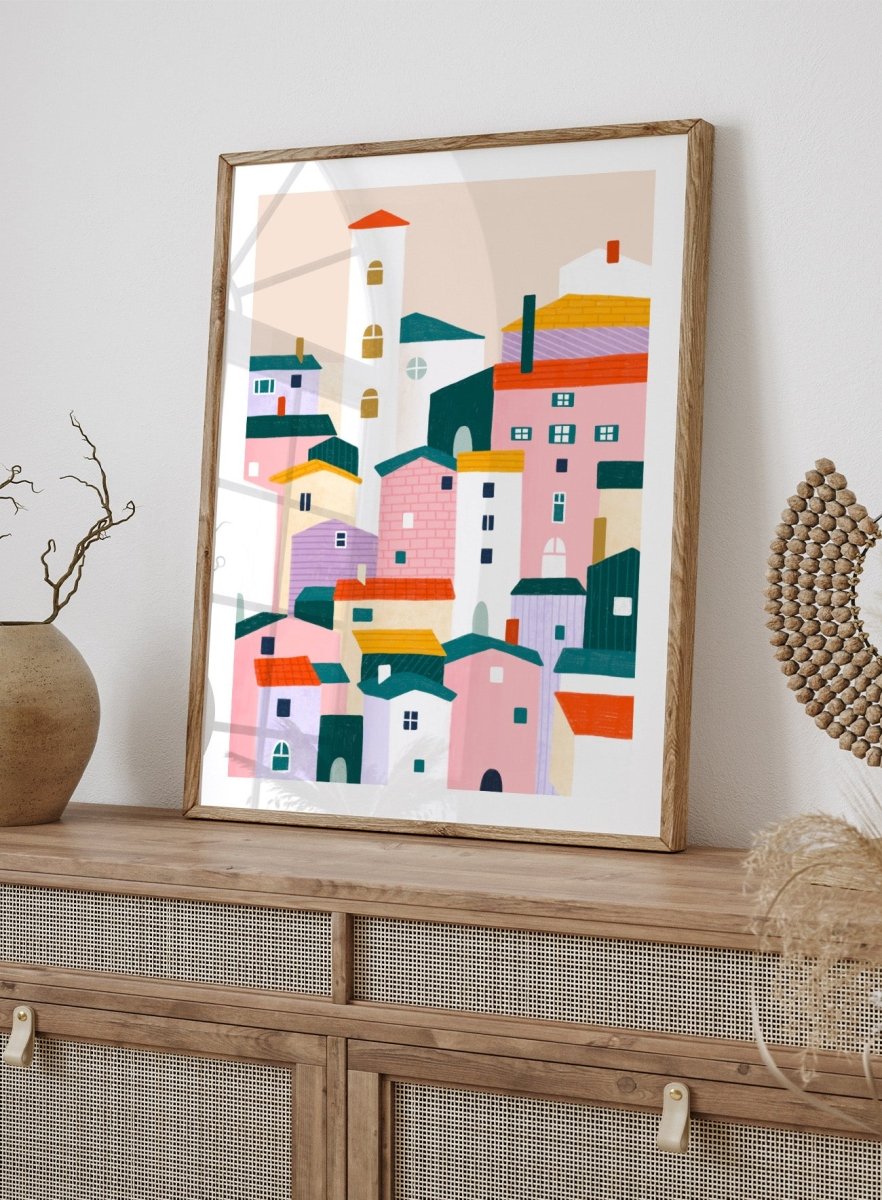 A framed minimalist art print depicting a cluster of stylized pastel houses and a church tower in pink, peach, teal, yellow, and cream tones. Charming Colorful Houses by Bea Müller premium framed fine art print featuring a playful town of pink, lavender, cream, and teal buildings.