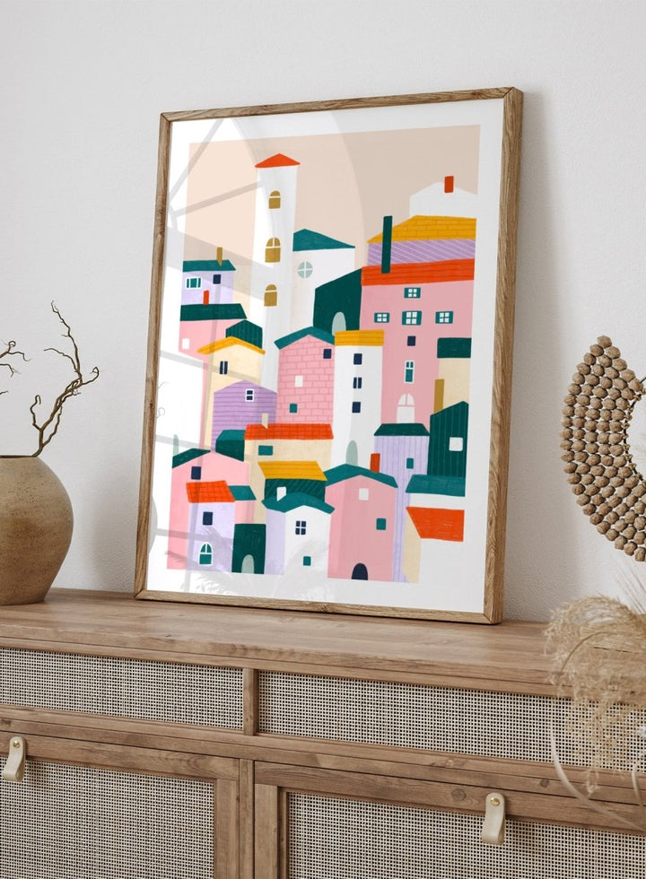 A framed minimalist art print depicting a cluster of stylized pastel houses and a church tower in pink, peach, teal, yellow, and cream tones. Charming Colorful Houses by Bea Müller premium framed fine art print featuring a playful town of pink, lavender, cream, and teal buildings.