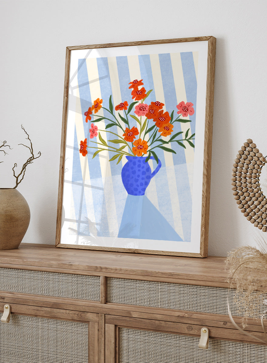 A framed art print featuring a stylized illustration of a blue patterned pitcher vase filled with orange, red, and pink flowers against a blue and white striped background. Cheerful Floral Pitcher by Bea Müller floral still life art print featuring orange and pink flowers in a bright blue jug with striped blue decor.