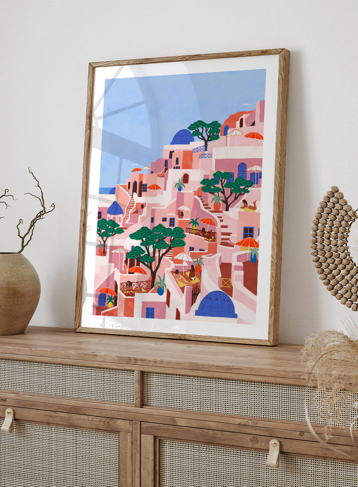 A stylized art print depicting a hillside Mediterranean village with pink and peach buildings, blue domes, green trees, and a light blue sky. Santorini Summer Dream by Ceyda Alasar art print featuring blue domes, terraced Mediterranean architecture, orange umbrellas, and sunny Greek island charm.