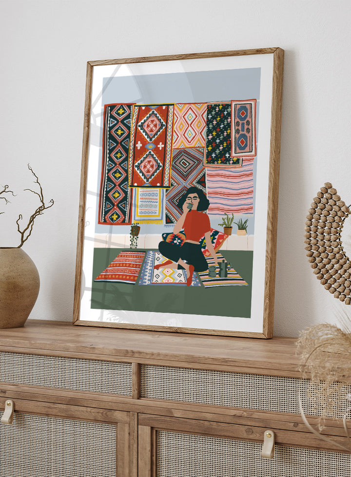 A framed art print featuring a stylized illustration of a woman sitting cross-legged on a patterned rug, surrounded by colorful geometric textiles hanging on a wall. Bohemian Coffee Morning by Ceyda Alasar interior art print featuring patterned rugs, floor cushions, potted plants, coffee, and colorful collected decor.