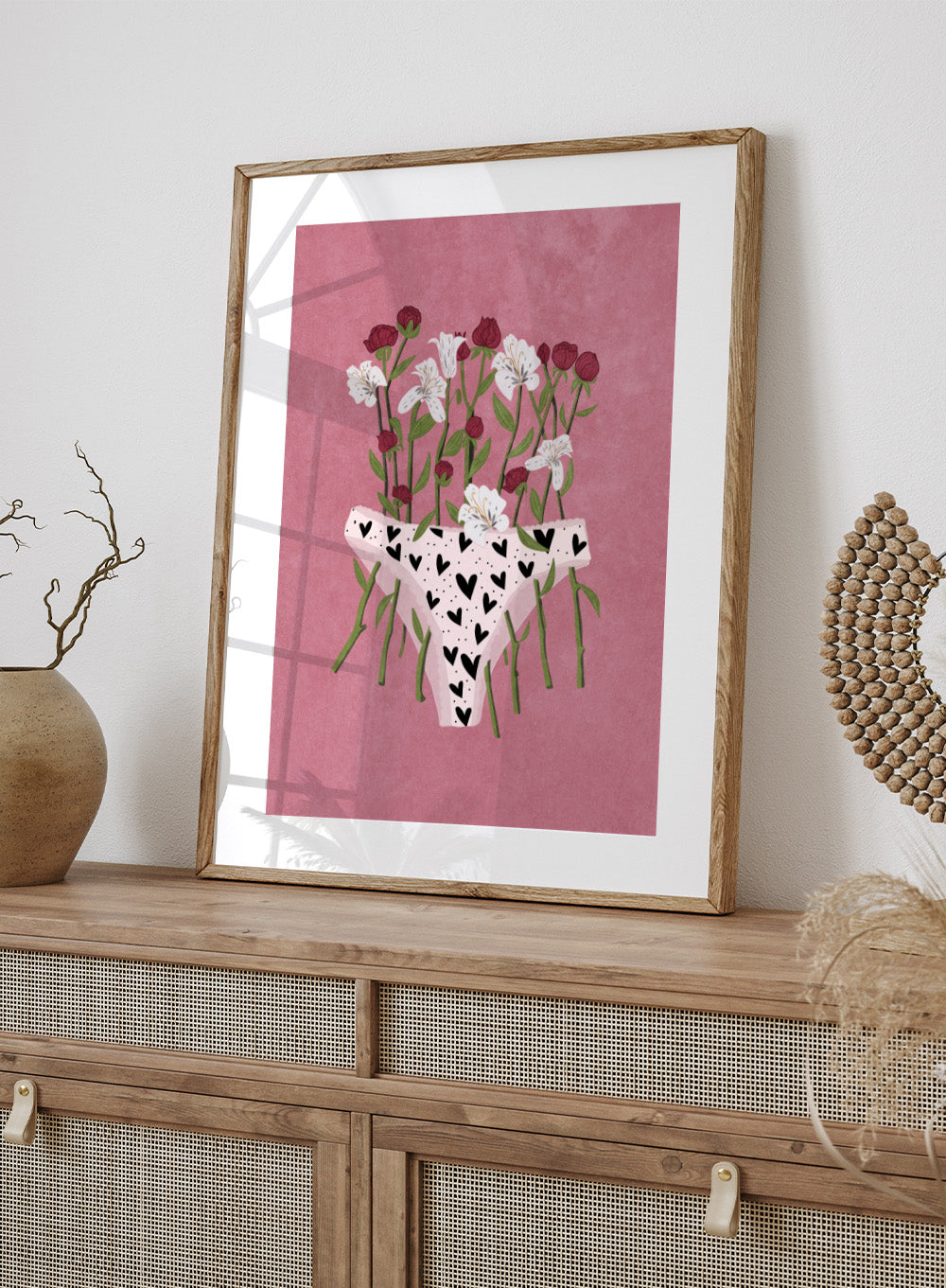 White underwear with black heart patterns surrounded by flowers on a pink background. Blooming Slip by Raissa Oltmanns features a feminist illustration of flowers growing from heart-patterned underwear on a pink background. Bold floral art print with strong feminine symbolism.