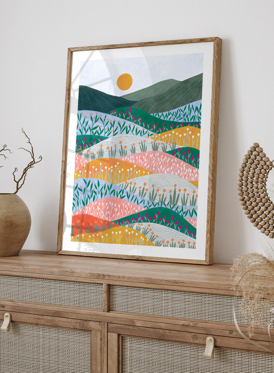 A framed art print featuring a stylized landscape illustration with rolling hills, wildflowers, mountains, and a sun in pastel and bright colors. Colorful Flower Hills by Ceyda Alasar botanical landscape art print featuring layered wildflower fields, rolling hills, mountains, and a golden sun.