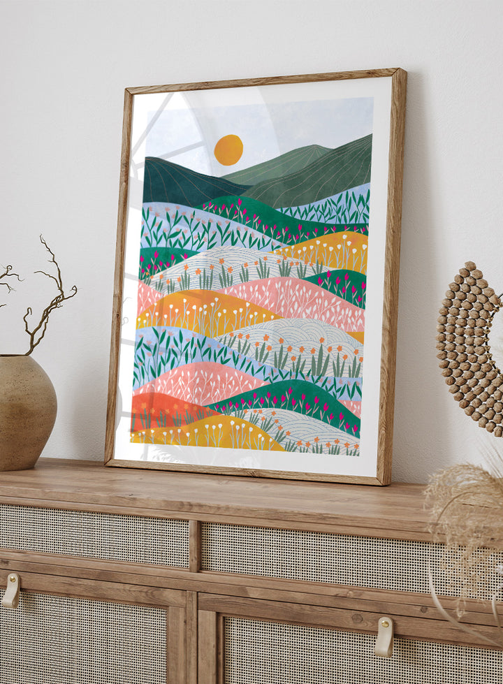 A framed art print featuring a stylized landscape illustration with rolling hills, wildflowers, mountains, and a sun in pastel and bright colors. Colorful Flower Hills by Ceyda Alasar botanical landscape art print featuring layered wildflower fields, rolling hills, mountains, and a golden sun.