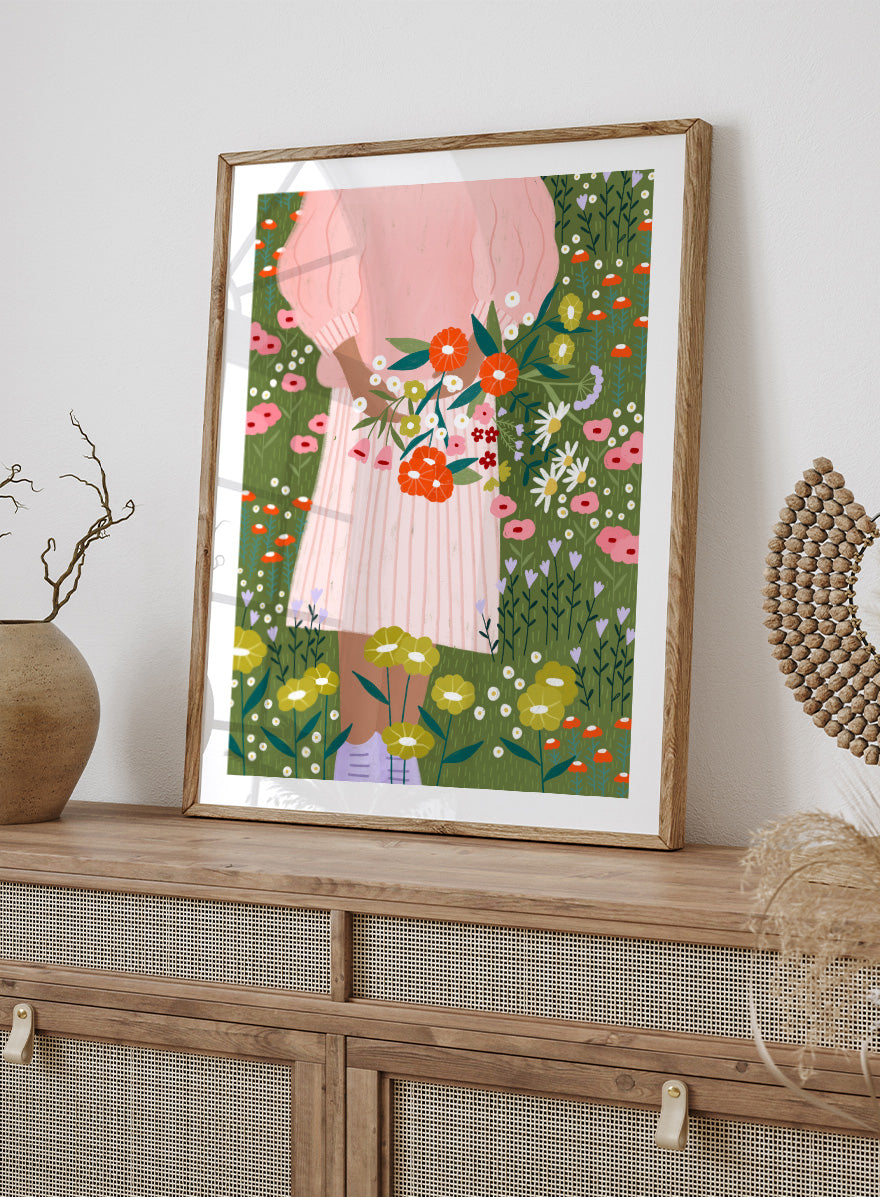A framed art print featuring a stylized illustration of a person in a pink striped dress holding a bouquet of orange, red, and white flowers in a green field of wildflowers. Secret Garden Bouquet by Bea Müller floral art print featuring a woman gathering wildflowers in a colorful garden filled with poppies, daisies, and greenery.