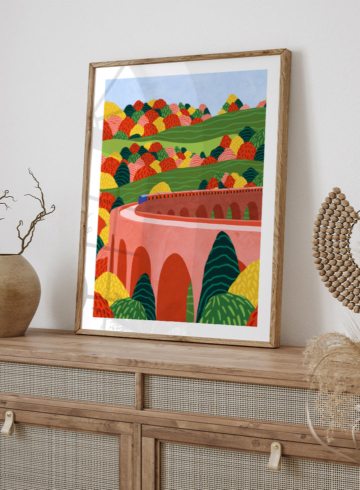 A framed art print featuring a stylized landscape illustration with rolling green hills, colorful patterned trees, and a pink arch bridge under a light blue sky. Winding Autumn Viaduct by Ceyda Alasar travel art print featuring a colorful train, arched bridge, rolling hills, and bold autumn foliage.