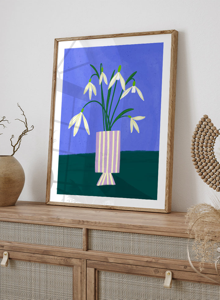 A framed art print featuring a stylized illustration of white snowdrops with green stems in a lavender and cream striped vase, set against a blue and dark green background. Snowdrops in Striped Vase by Bea Müller fine art print featuring white snowdrops, a lilac striped vase, and a deep blue background.