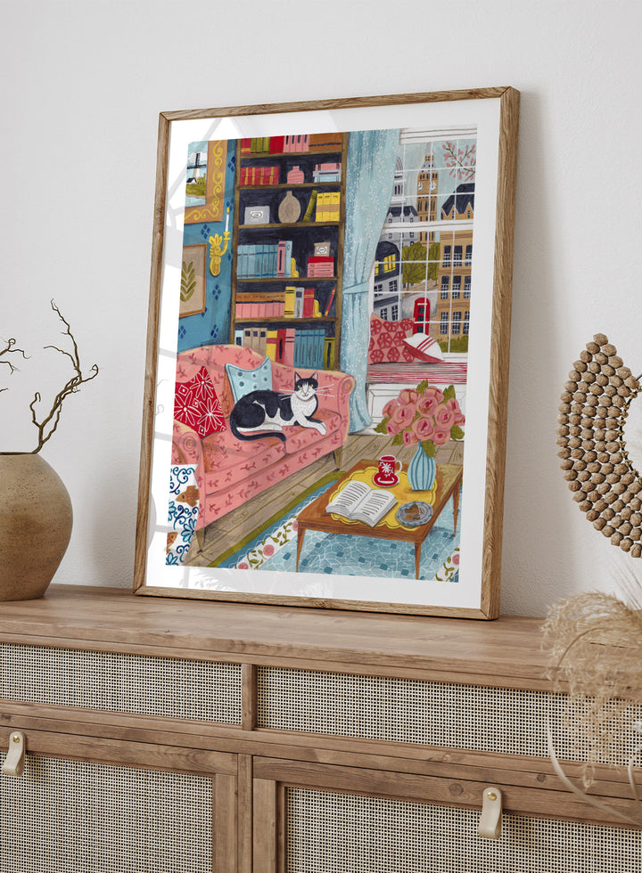 Illustration of a cozy living room with a cat on a pink sofa, bookshelf, and coffee table. Cozy London Cat by Caroline Bonne Müller is a fine art print and wall poster featuring a cat lounging in a colorful living room with a London city view.
