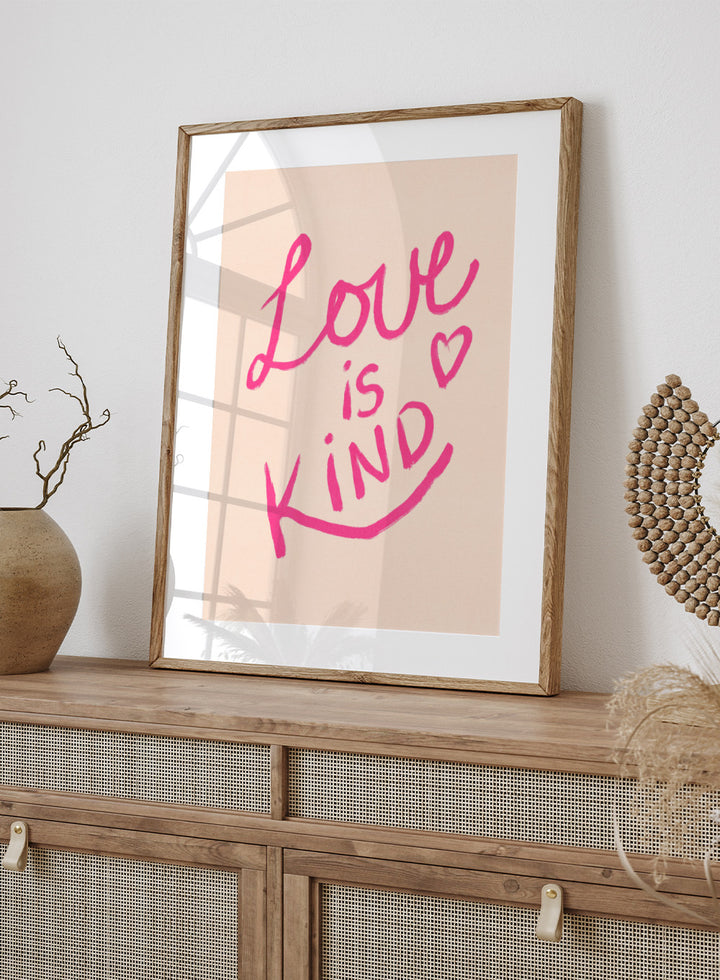 Pink text 'Love is Kind' with a heart on a light pink background. Love Is Kind by Dirkie Nell is a pink typography art print with handwritten lettering and heart detail. A joyful modern love wall poster.