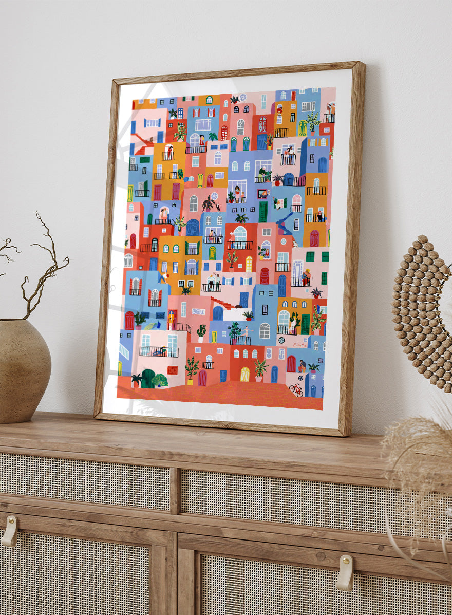 A framed art print featuring a colorful, whimsical illustration of Mediterranean-style buildings with people, animals, and plants. Colorful Town Life by Ceyda Alasar city art print featuring vibrant buildings, balconies, neighbors, plants, and lively urban details in bold color.