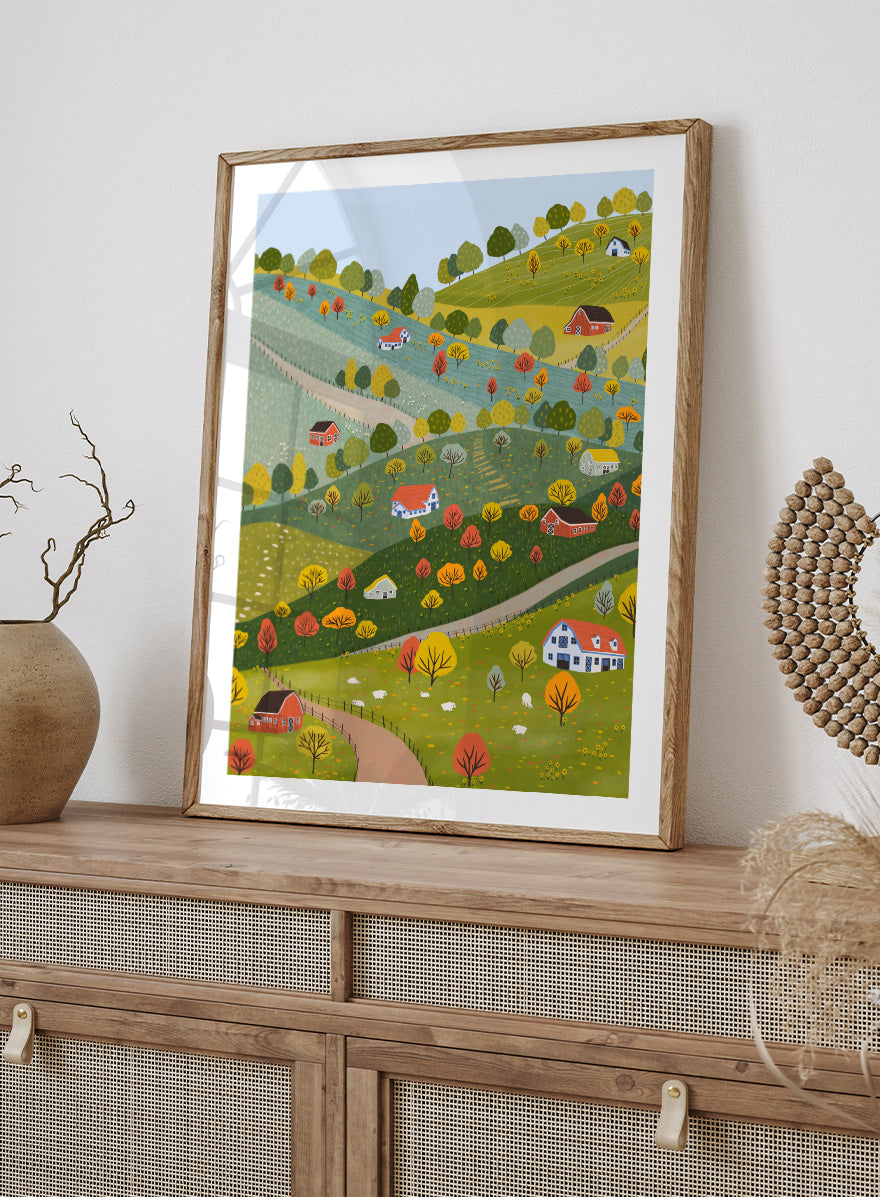 A framed art print featuring a whimsical, stylized illustration of a rolling green and yellow countryside landscape with red houses, colorful autumn trees, winding roads, and a pale blue sky. Vibrant Autumn Village Hills by Ceyda Alasar art print with rolling countryside, colorful fall trees, farmhouses, barns, sheep, and winding roads.
