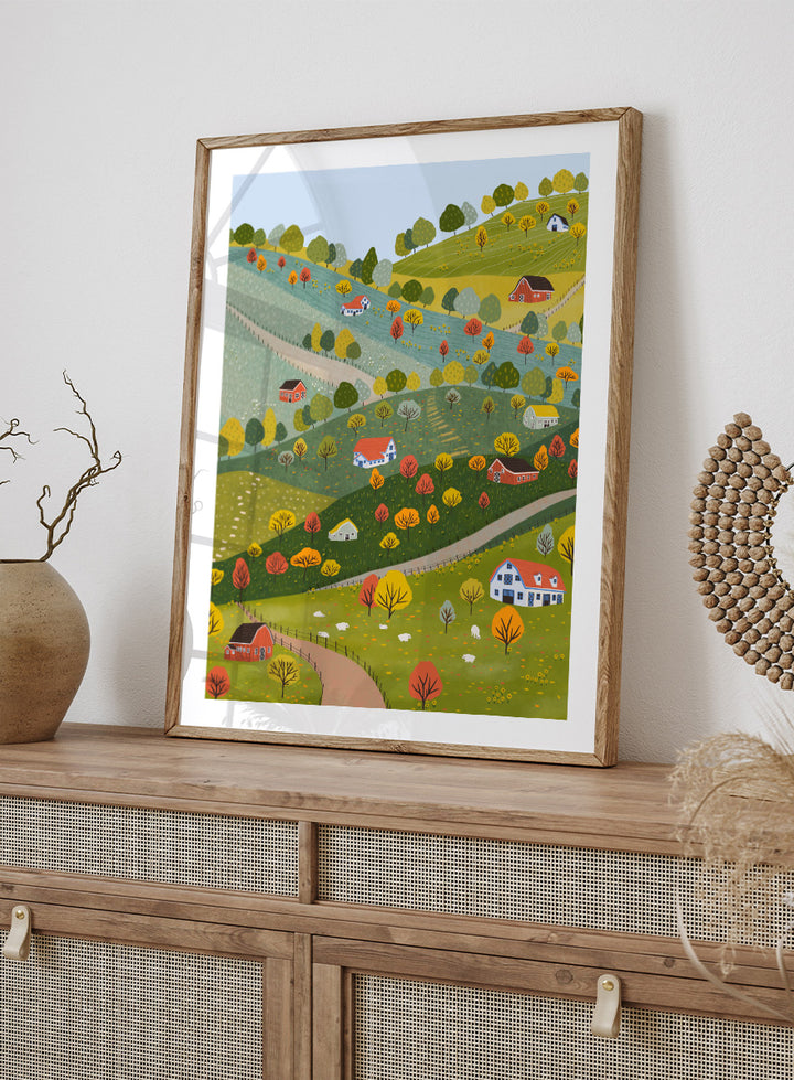 A framed art print featuring a whimsical, stylized illustration of a rolling green and yellow countryside landscape with red houses, colorful autumn trees, winding roads, and a pale blue sky. Vibrant Autumn Village Hills by Ceyda Alasar art print with rolling countryside, colorful fall trees, farmhouses, barns, sheep, and winding roads.