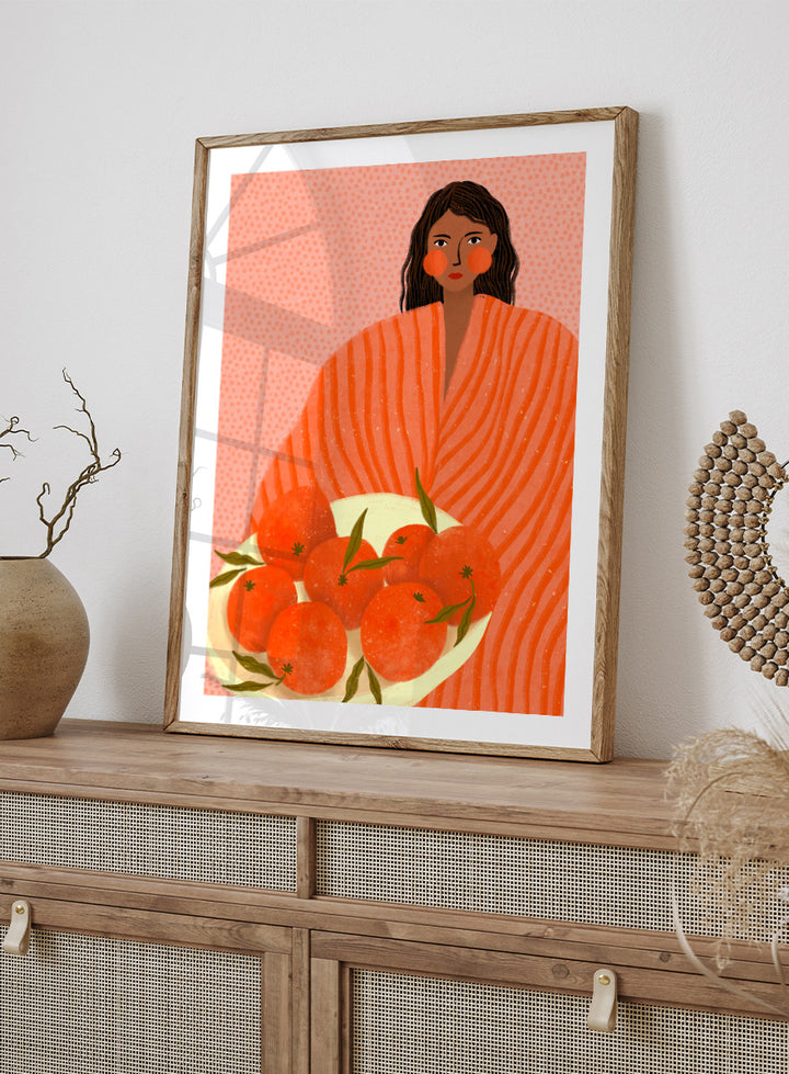 A stylized art print featuring a woman with dark hair wrapped in a bright orange striped blanket, holding a white plate of oranges against a pink dotted background. Woman Holding Oranges by Bea Müller colorful kitchen art print featuring a woman carrying fresh citrus fruit in warm orange and coral tones.