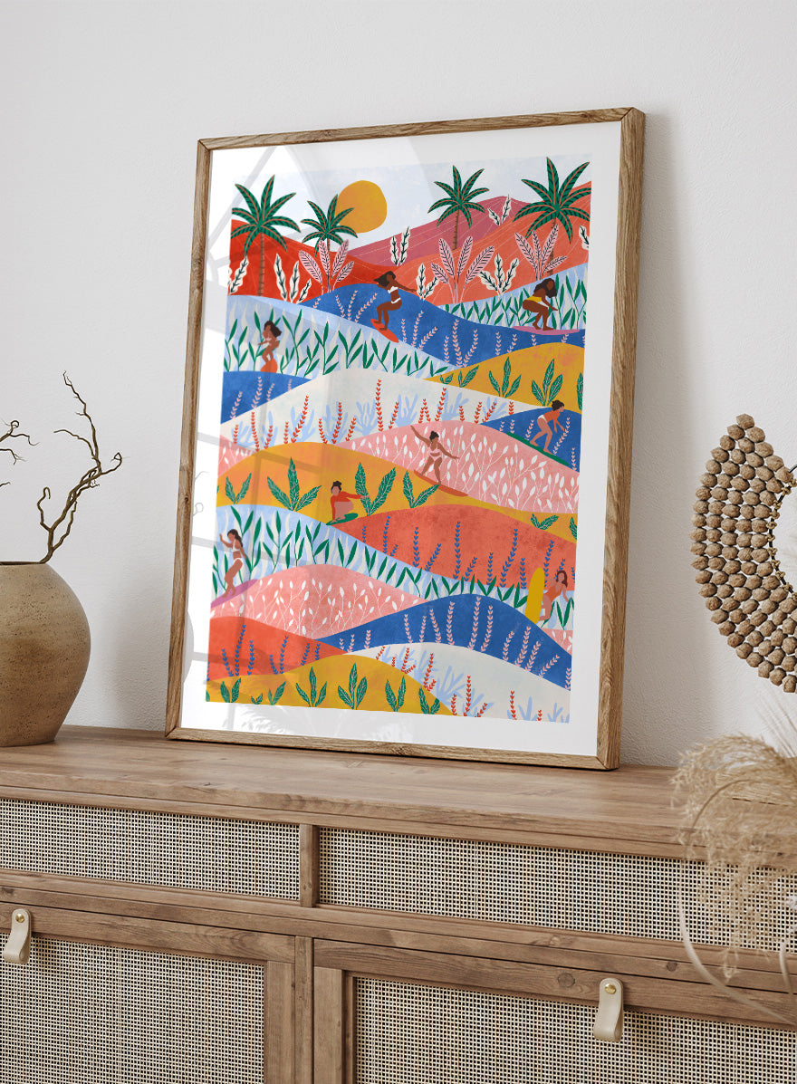 A framed art print featuring a colorful, stylized landscape illustration with rolling hills, palm trees, a sun, and small human figures in a folk-art style. Tropical Surf Girls by Ceyda Alasar surf art print featuring women riding waves through a colorful palm-filled landscape in bold tropical tones.